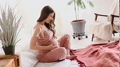 young pregnant woman with long hair in pajamas examines baby clothes for newborns while sitting on bed. mother expecting child. Preparing for childbirth pregnant woman stroking her belly