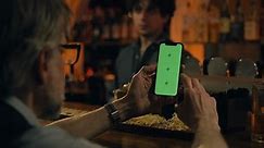 Mature man uses mobile phone with green screen, bets on football match online or surfs the Internet. Sports fan watches bookmaker ratings, talks with bartender sitting at bar counter in pub. Mock-up.