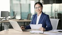 Portrait of happy asian female financier working on a laptop doing paperwork sitting at workplace at desk in a business office. Smiling accountant in a formal suit in glasses posing looking at camera