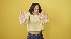 Panicked, young hispanic woman, a beautiful portrait of fear and shock. terrified expression, hands up in stop gesture, shouting, isolated on yellow background. sweater-wearing fright.