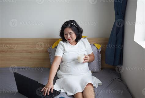 Happy Young Asian Pregnant Woman drinking milk on bed at home while holding her belly and ...