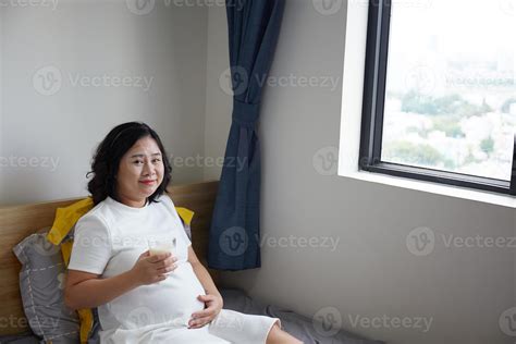 Happy Young Asian Pregnant Woman drinking milk on bed at home while holding her belly and ...