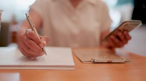 Download Cafe Note Taking. Close-up of hands meticulously writing on a notepad, a smartphone rests nearby, suggesting a blend of digital and analog work in a cafe setting. for free