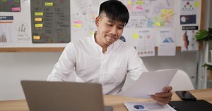 Portrait of Exhausted asian man typing laptop and checking document while sitting at workplace desk in office. Focused young guy working on project document at modern office.
