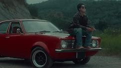 A brutal girl in a brown leather jacket sits on the hood of her vintage red car and smokes