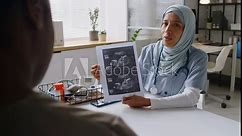 Medium over-shoulder shot of young female ob-gyn in Islamic hijab sitting at desk in clinic or hospital, showing ultrasound image of foetus in womb to pregnant African American patient, while talking