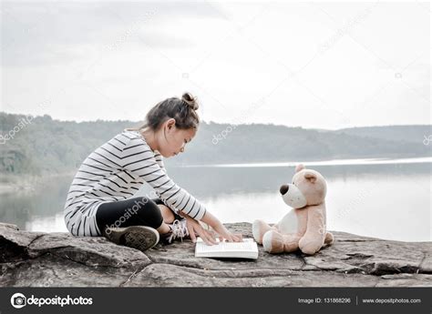 Chica leyendo un libro de elizabeth eulberg; Asiática chica leyendo un libro con osito de peluche en la ...